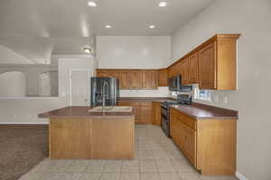 Kitchen with double oven range, refrigerator with ice dispenser, a kitchen island with sink, light tile patterned floors, and dark countertops
