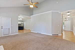 Unfurnished living room with light colored carpet, high vaulted ceiling, a chandelier, ceiling fan, and crown molding