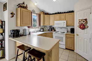 Kitchen featuring white appliances, light brown cabinetry, lofted ceiling, a kitchen breakfast bar, and a peninsula
