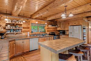 Kitchen with stainless steel appliances, log walls, tile counters, a wooden ceiling with exposed beams, and light wood floors