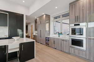 Kitchen featuring modern cabinets, stainless steel double oven, light stone countertops, light wood finished floors, and wine cooler