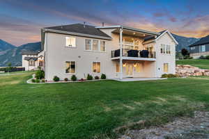 Back of property at dusk featuring a balcony, a patio, stucco siding, a mountain view, and a lawn