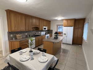 Kitchen featuring white appliances, light tile patterned floors, a center island, tasteful backsplash, and brown cabinets