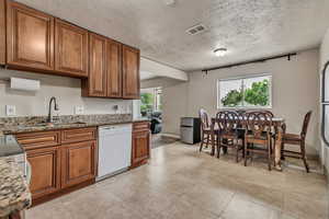 Kitchen with dishwasher, brown cabinets, a textured ceiling, stone countertops, and light tile patterned floors