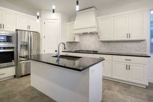 Gourmet kitchen with stainless steel appliances, custom range hood, black quartz countertops, tasteful backsplash, white cabinets, and vaulted ceiling.