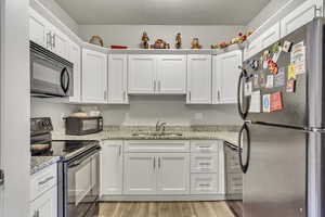 Kitchen with black appliances, a sink, white cabinets, light wood-type flooring, and light stone countertops