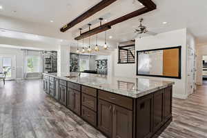 Kitchen featuring light wood finished floors, dark brown cabinetry, a ceiling fan, beamed ceiling, and light stone countertops