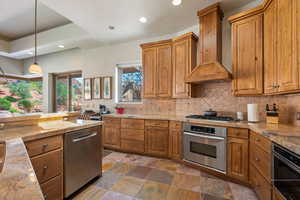 Kitchen featuring stainless steel appliances, premium range hood, brown cabinetry, decorative backsplash, and recessed lighting