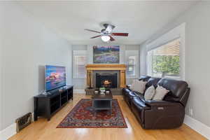 Living room featuring wood finished floors, ceiling fan, and a warm lit fireplace