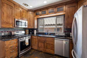 Kitchen featuring appliances with stainless steel finishes, tasteful backsplash, brown cabinetry, glass insert cabinets, and dark stone finish floors