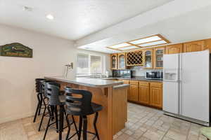 Kitchen featuring white refrigerator with ice dispenser, stainless steel microwave, a kitchen bar, a peninsula, and glass insert cabinets