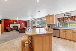 Kitchen with dishwasher, light brown cabinetry, light countertops, a glass covered fireplace, and a center island