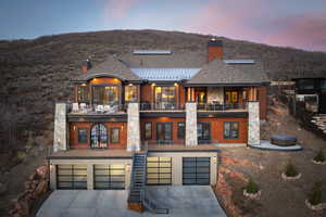 Back of property at dusk with a balcony, a chimney, a garage, driveway, and stone siding