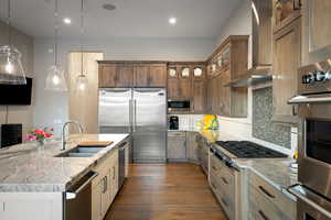 Kitchen featuring built in appliances, wall chimney range hood, decorative backsplash, light stone countertops, and recessed lighting