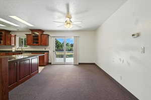 Kitchen with dark countertops, a ceiling fan, glass insert cabinets, light colored carpet, and plenty of natural light
