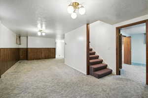 Finished basement featuring carpet flooring, stairway, a textured ceiling, a wainscoted wall, and wood walls