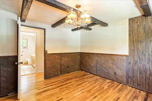 Empty room with beamed ceiling, light wood-type flooring, a wainscoted wall, a chandelier, and a textured ceiling