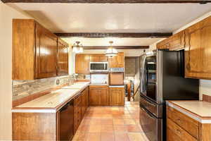 Kitchen featuring appliances with stainless steel finishes, tile counters, brown cabinets, tasteful backsplash, and beamed ceiling