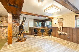 Indoor dry bar with beam ceiling, a wainscoted wall, plenty of natural light, wood finished floors, and wooden walls