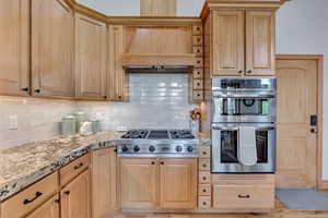 Kitchen with stainless steel appliances, custom range hood, backsplash, light stone countertops, and light brown cabinets