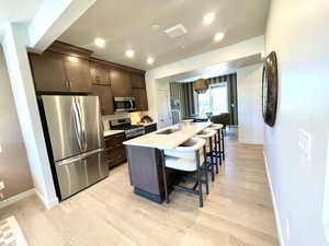 Kitchen featuring appliances with stainless steel finishes, dark brown cabinets, light wood-style floors, a kitchen bar, and a center island with sink