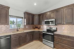 Kitchen with appliances with stainless steel finishes, vaulted ceiling, light wood-style floors, dark brown cabinets, and light stone countertops