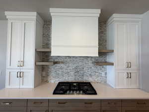Kitchen with open shelves, stainless steel gas stovetop, backsplash, light stone countertops, and range hood