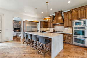 Kitchen with built in appliances, custom exhaust hood, a center island with sink, brown cabinets, and recessed lighting