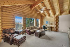 Living room with a wooden ceiling with exposed beams, log walls, carpet floors, ceiling fan, and high vaulted ceiling