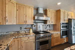 Kitchen featuring wall chimney range hood, stainless steel appliances, light brown cabinets, a textured ceiling, and dark wood finished floors