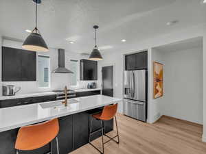 Kitchen with stainless steel refrigerator with ice dispenser, wall chimney range hood, dark cabinetry, light countertops, and decorative light fixtures
