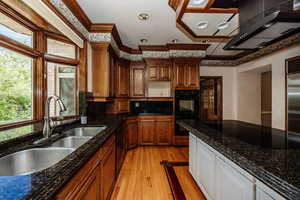 Kitchen featuring black appliances, range hood, light wood-type flooring, crown molding, and dark stone countertops