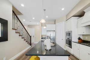 Kitchen featuring stainless steel appliances, premium range hood, an island with sink, wood finished floors, and white cabinets
