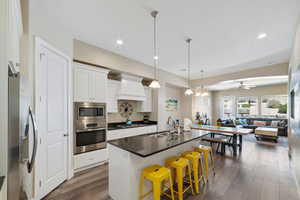 Kitchen with stainless steel appliances, custom exhaust hood, dark countertops, open floor plan, and dark wood-style floors