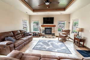 Living room with a raised ceiling, a ceiling fan, ornamental molding, a tiled fireplace, and stone tile flooring