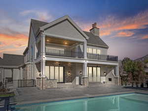 Back of house at dusk with a patio area, a chimney, an outdoor pool, a balcony, and stone siding