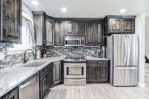 Kitchen with appliances with stainless steel finishes, light stone countertops, light wood-style flooring, decorative backsplash, and dark brown cabinetry