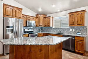 Kitchen featuring stainless steel appliances, lofted ceiling, decorative backsplash, a kitchen island, and brown cabinetry