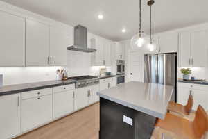 Kitchen featuring wall chimney exhaust hood, stainless steel appliances, tasteful backsplash, dark countertops, and pendant lighting