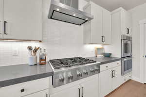 Kitchen with wall chimney range hood, appliances with stainless steel finishes, dark countertops, decorative backsplash, and white cabinets