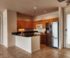 Kitchen featuring white appliances, dark countertops, a peninsula, brown cabinetry, and recessed lighting