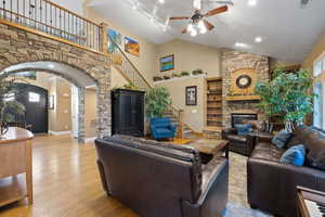Living room with stairway, a stone fireplace, a ceiling fan, wood finished floors, and high vaulted ceiling