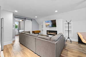 Living room featuring light wood-style floors, a barn door, vaulted ceiling, a glass covered fireplace, and built in features