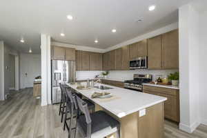 Kitchen featuring stainless steel appliances, light wood-style floors, a kitchen bar, a center island with sink, and light countertops