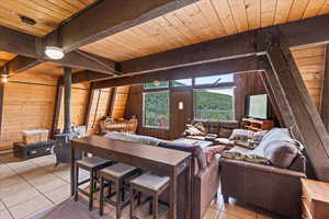 Living room with wood walls, light tile patterned floors, a wooden ceiling with exposed beams, and a wood stove