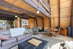 Living room with a wood stove, light tile patterned floors, wooden walls, and a wooden ceiling with exposed beams
