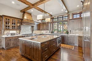 Kitchen featuring built in appliances, custom exhaust hood, a center island with sink, beamed ceiling, and dark wood-style flooring