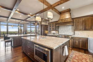 Kitchen featuring appliances with stainless steel finishes, coffered ceiling, an island with sink, custom exhaust hood, and backsplash