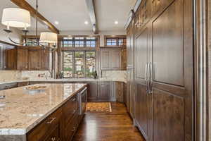 Kitchen with beamed ceiling, built in appliances, dark wood-type flooring, tasteful backsplash, and light stone counters