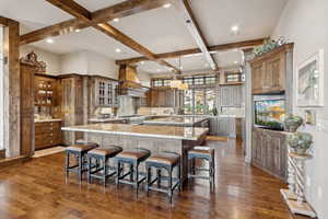 Kitchen with a large island with sink, beam ceiling, dark wood finished floors, glass insert cabinets, and light stone countertops